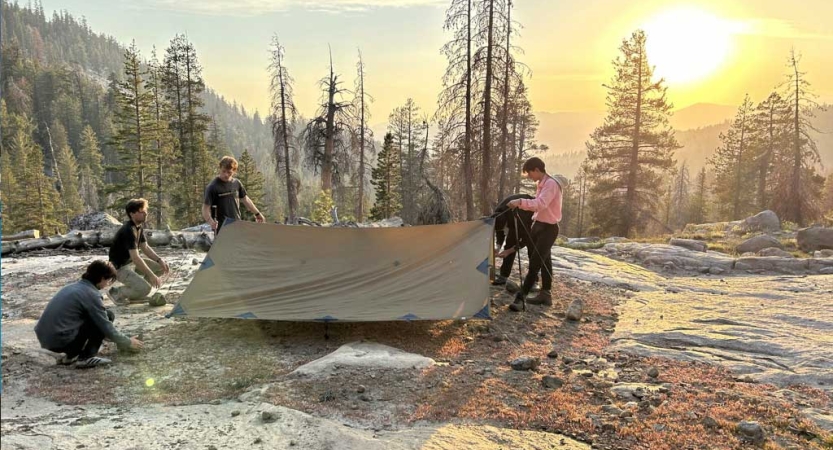 A group of people set up a tarp shelter in a mountainous landscape with evergreen trees. 